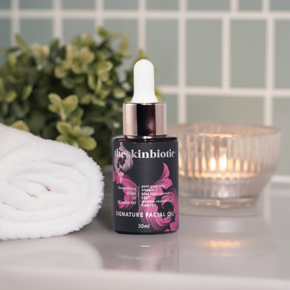 theSkinBiotic Signature Facial Oil  picture of a bottle with a dropper on a bathroom bench with gentle candle lighting, a plant and white towel